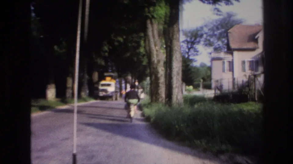 8mm home movie frame, France 1967: male cyclist riding away from camera on a plane-tree-lined road with a yellow van ahead.