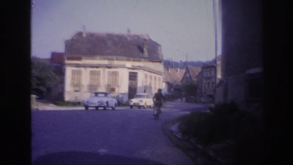 8mm home movie frame, France 1967: village center with large multi-story stone building, two white compact cars parked, cyclist figure in foreground.