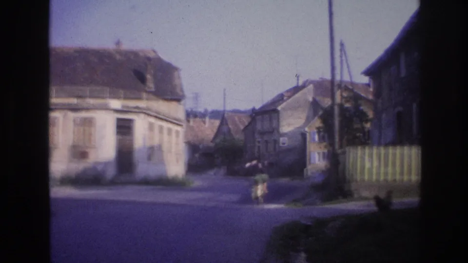 8mm frame, France 1967: stone village buildings from moving vehicle POV, vertical wooden fence on right, figure visible in road.
