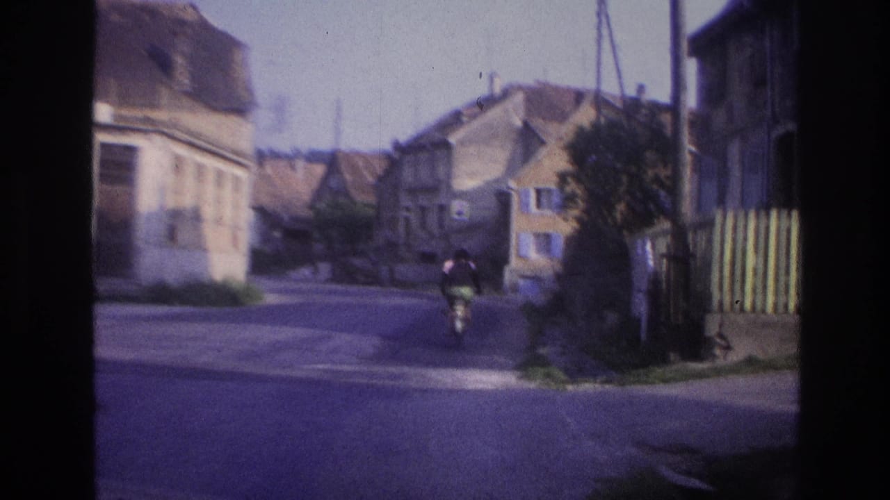 Cyclist moving away from camera down a narrow European street lined with stone and stucco gabled buildings, captured on 8mm film described as France circa 1967.