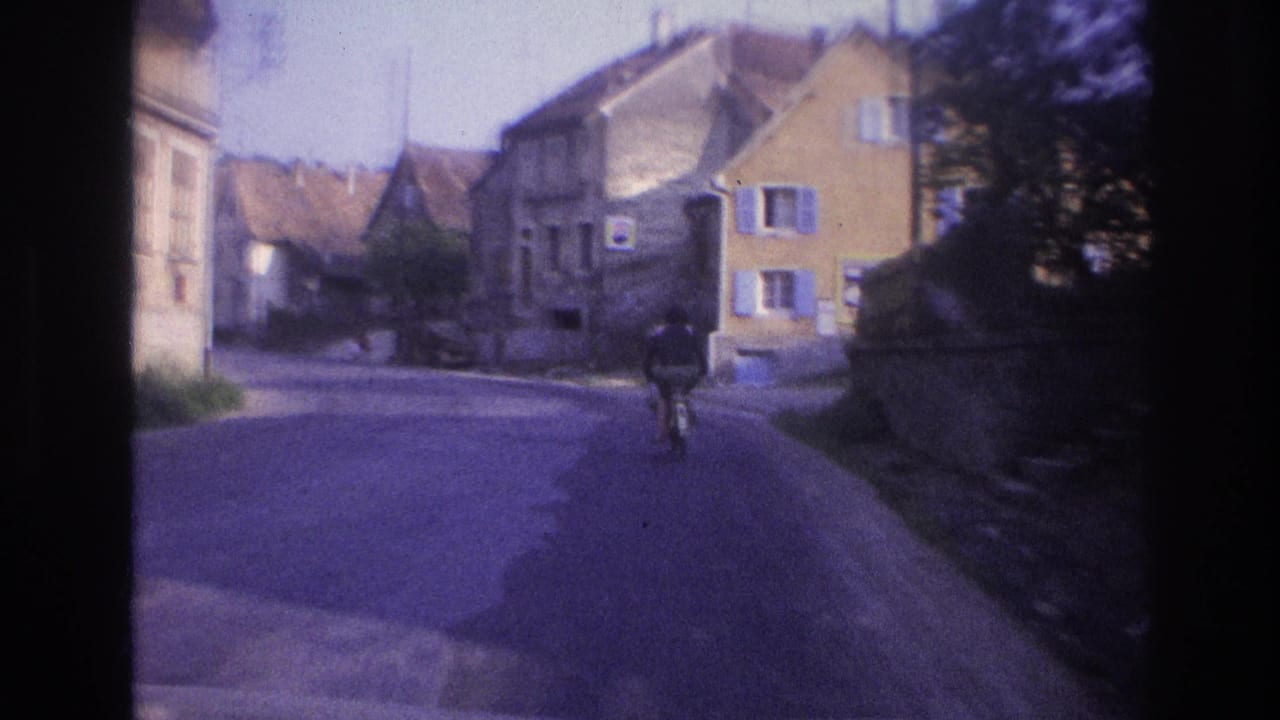Cyclist advancing along a small-town street with stone buildings, blue painted shutters, and tiled roofs; 8mm archival footage described as France circa 1967.