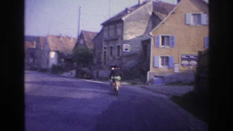 Touring cyclist with rear panniers riding past a prominent ochre-coloured building with blue shutters and street-level notices; 8mm scene described as France circa 1967.