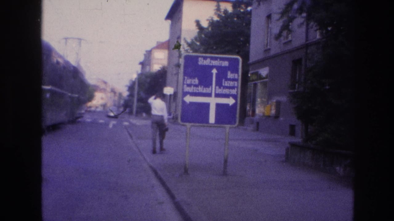 Man walking past a blue cross-shaped sign reading Stadtzentrum, Zürich and Bern at a late-1960s Central European tram intersection; violet 8mm color cast.