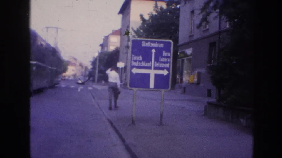 Man walking past a blue cross-shaped sign reading Stadtzentrum, Zürich and Bern at a late-1960s Central European tram intersection; violet 8mm color cast.