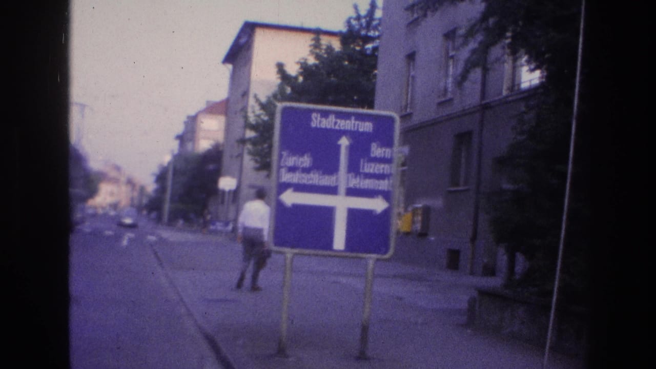 Man in white shirt and dark trousers near the blue directional sign; European apartment buildings and empty street visible behind him.