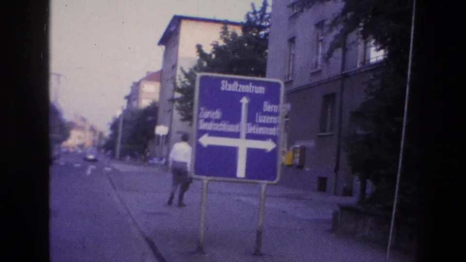 Man at mid-distance near the blue Stadtzentrum directional sign, European mid-century apartment blocks behind; late-1960s Central European city, 8mm violet cast.