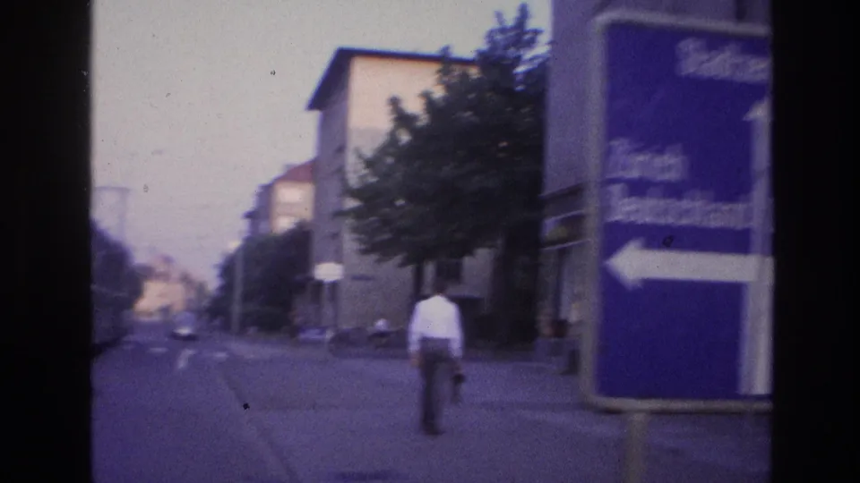 Close angle on the blue German-language road sign (Zürich left arrow, Stadtzentrum visible); man in white shirt walking away in background; street trees and European buildings frame the shot.