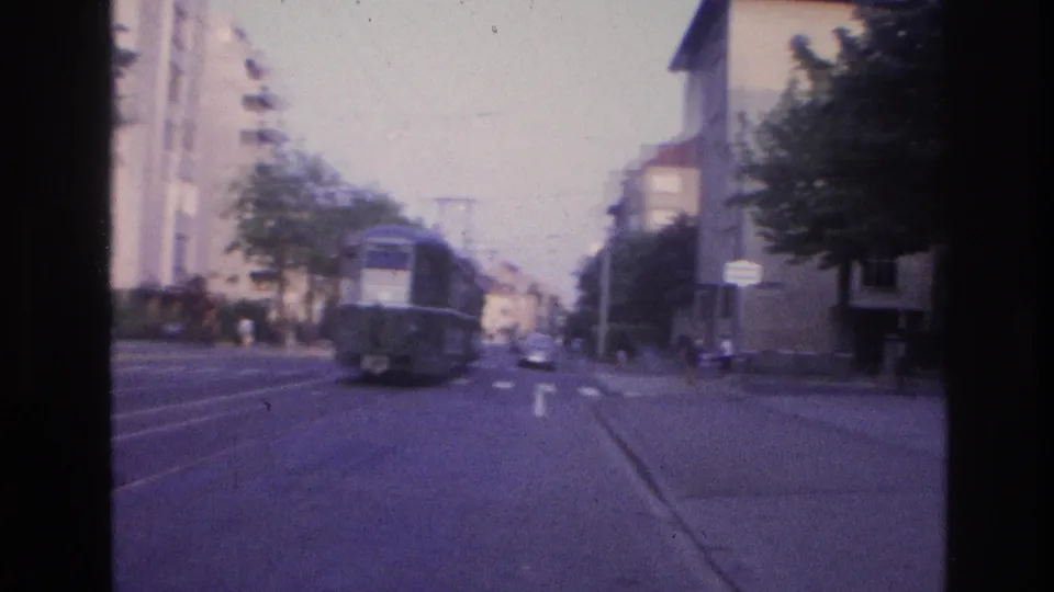 Tram on a Central European city street with mid-century apartment blocks, overhead tram wires and embedded tracks; blue directional sign partially visible at right; 8mm violet cast.