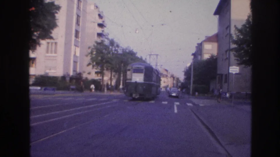 8mm film frame from France, circa 1967, showing a tram car on a residential boulevard flanked by apartment blocks and trees, with tram tracks and a zebra crossing in the foreground.