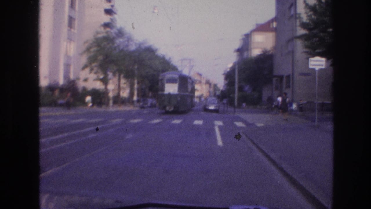 Tram moving toward an intersection on a residential boulevard with parked cars, trees, and a street sign visible on the right.
