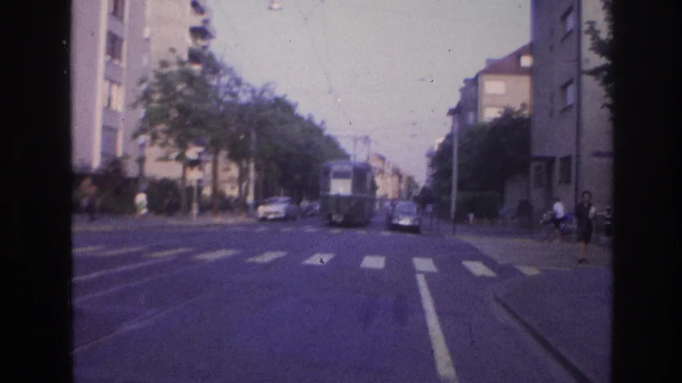 8mm frame from approximately 1967 France showing a pedestrian crossing near the camera vehicle, with tram tracks, parked 1960s-era cars, overhead wires, and residential apartment blocks on both sides.