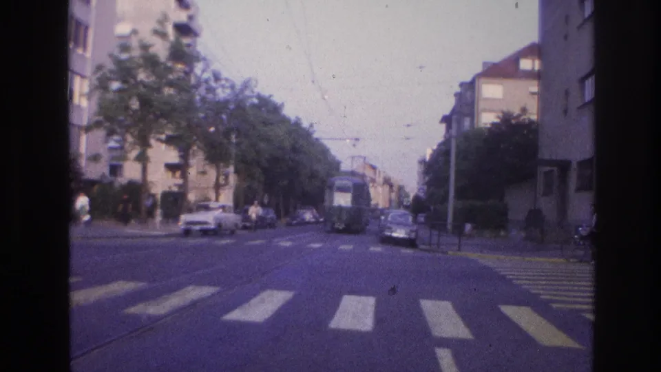 8mm frame from approximately 1967 France showing a boulevard intersection with 1960s-era cars, pedestrians, embedded tram tracks, and post-war apartment blocks under a pale sky.