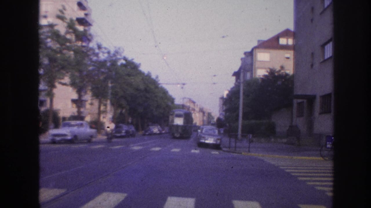 1960s French city street filmed from a moving vehicle showing overhead tram catenary, period saloon cars, a pedestrian crosswalk, and multi-story apartment buildings on both sides.
