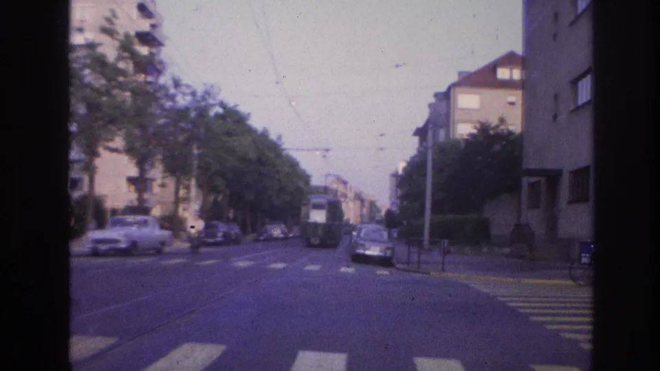 1960s French city street filmed from a moving vehicle showing overhead tram catenary, period saloon cars, a pedestrian crosswalk, and multi-story apartment buildings on both sides.