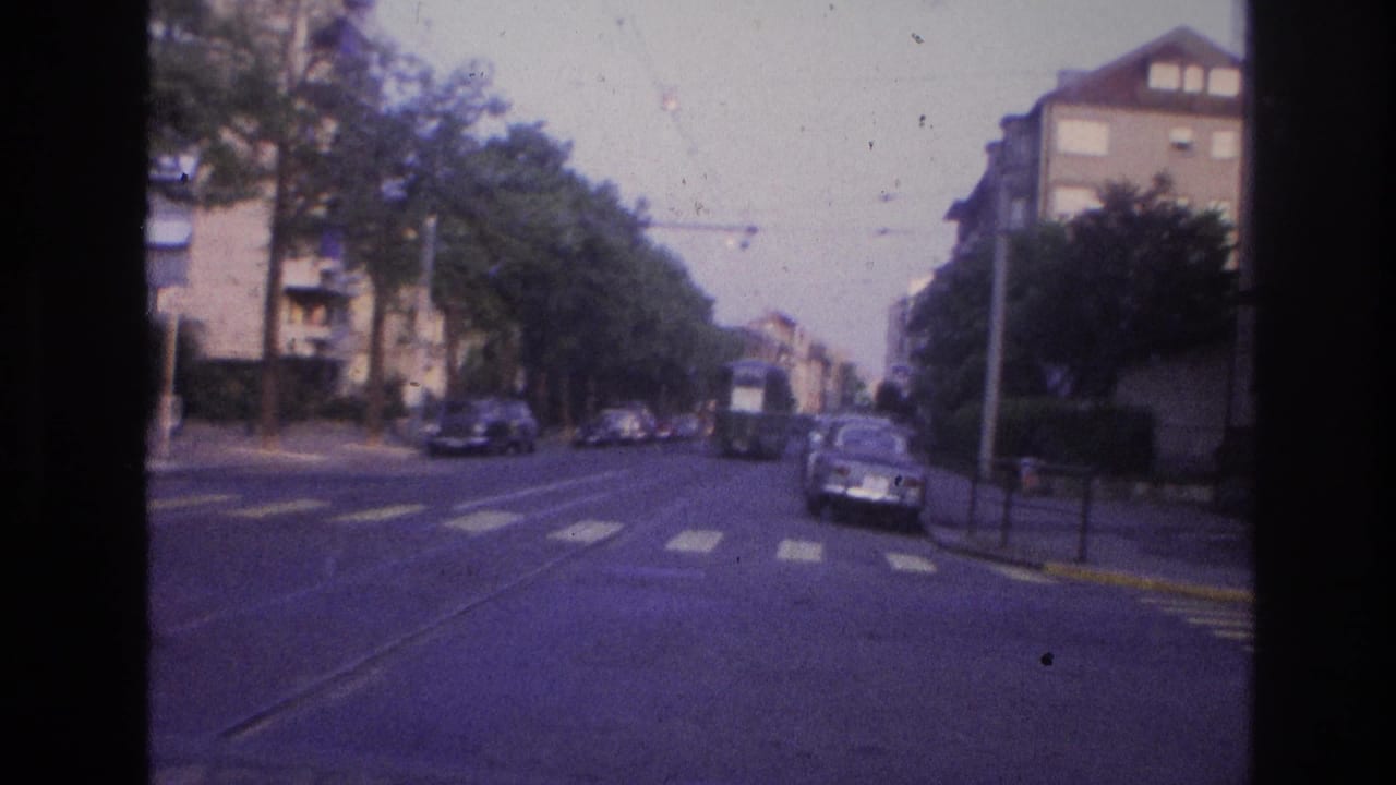 A European city intersection circa 1967 filmed from a moving vehicle, showing tram overhead lines, period cars, street trees, yellow road markings, and mid-century apartment buildings.