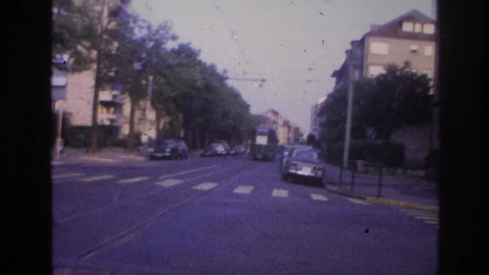 A European city intersection circa 1967 filmed from a moving vehicle, showing tram overhead lines, period cars, street trees, yellow road markings, and mid-century apartment buildings.