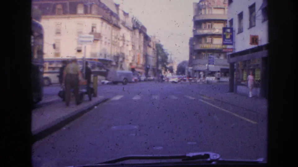1960s European commercial street filmed through a car windscreen, with ornate multi-story shopfronts, pedestrians on the pavement, directional road signs, and legible commercial signage including a large letter-R mark.