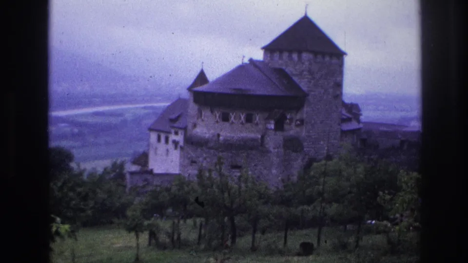 An unidentified medieval stone castle with multiple pointed towers and steep dark roofs, filmed on 8mm home movie in France circa 1967, overlooking a misty hillside valley under overcast skies.
