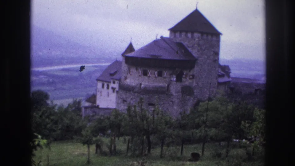 8mm home-movie frame from France, 1967, showing a stone castle with fortified towers above a broad valley, small dark shape visible mid-frame, overcast sky.