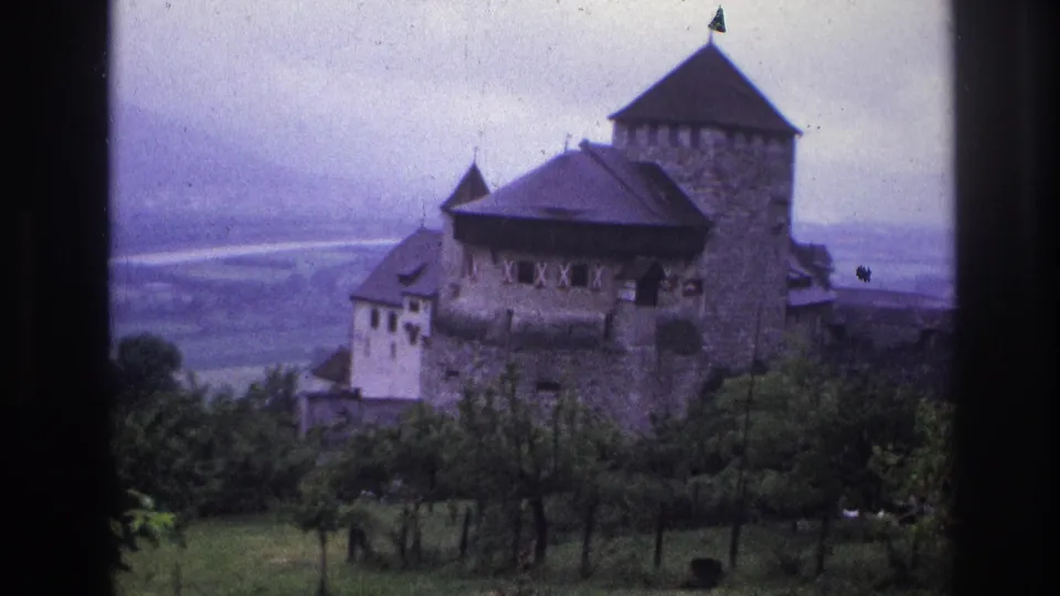 8mm home-movie frame circa 1967, France, showing a stone fortified castle with possible pennant on tower, green hillside, and broad hazy valley.