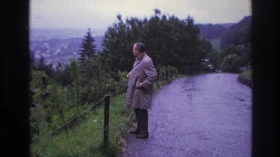 8mm home-movie frame from France, 1967, showing an adult male in a light coat on a winding road overlooking a broad valley, with wooden fence posts and dense green trees.