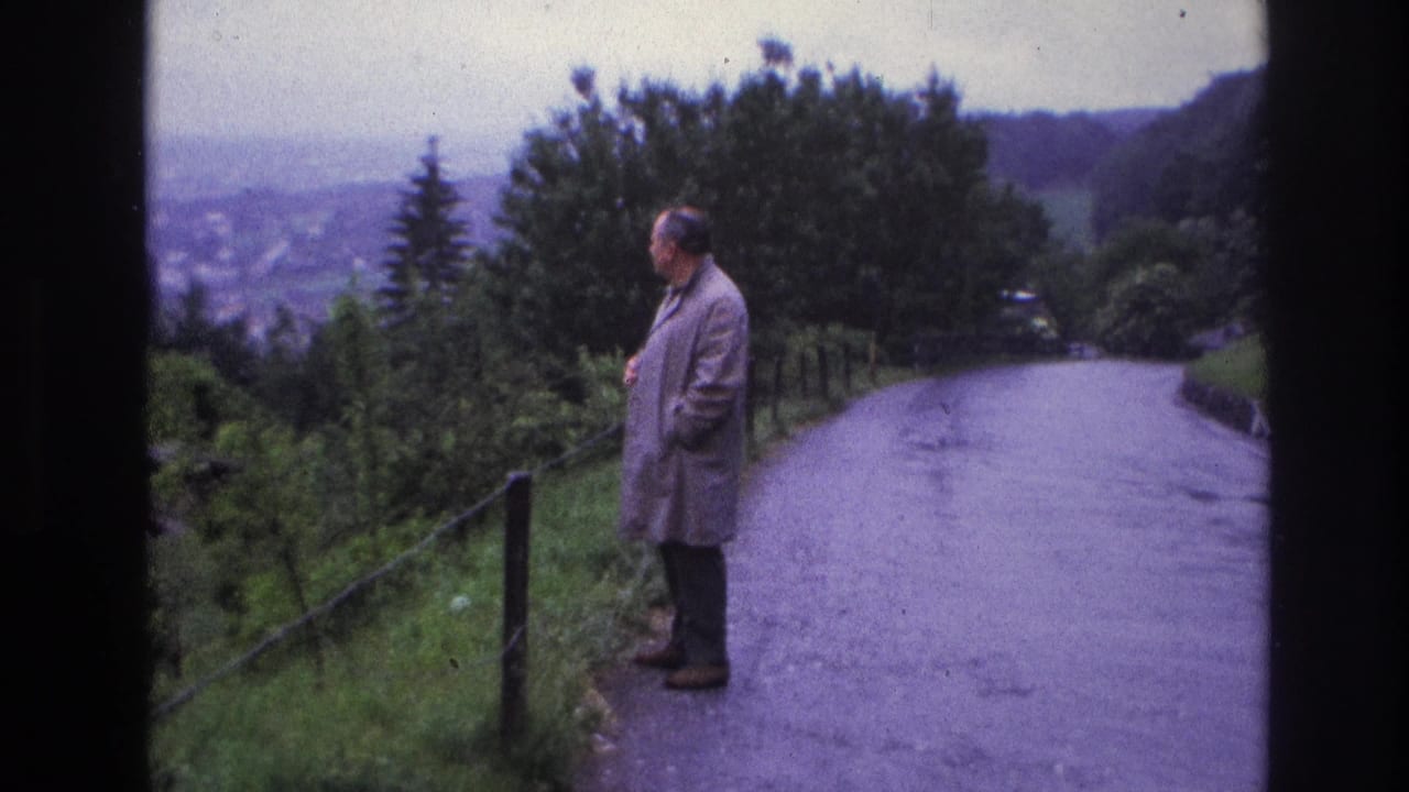 Middle-aged man in grey raincoat stands in profile on a hillside road, France, circa 1967; tree-lined slopes and a distant valley visible behind him.