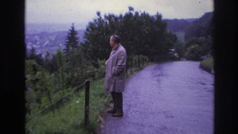 Middle-aged man in grey raincoat stands in profile on a hillside road, France, circa 1967; tree-lined slopes and a distant valley visible behind him.