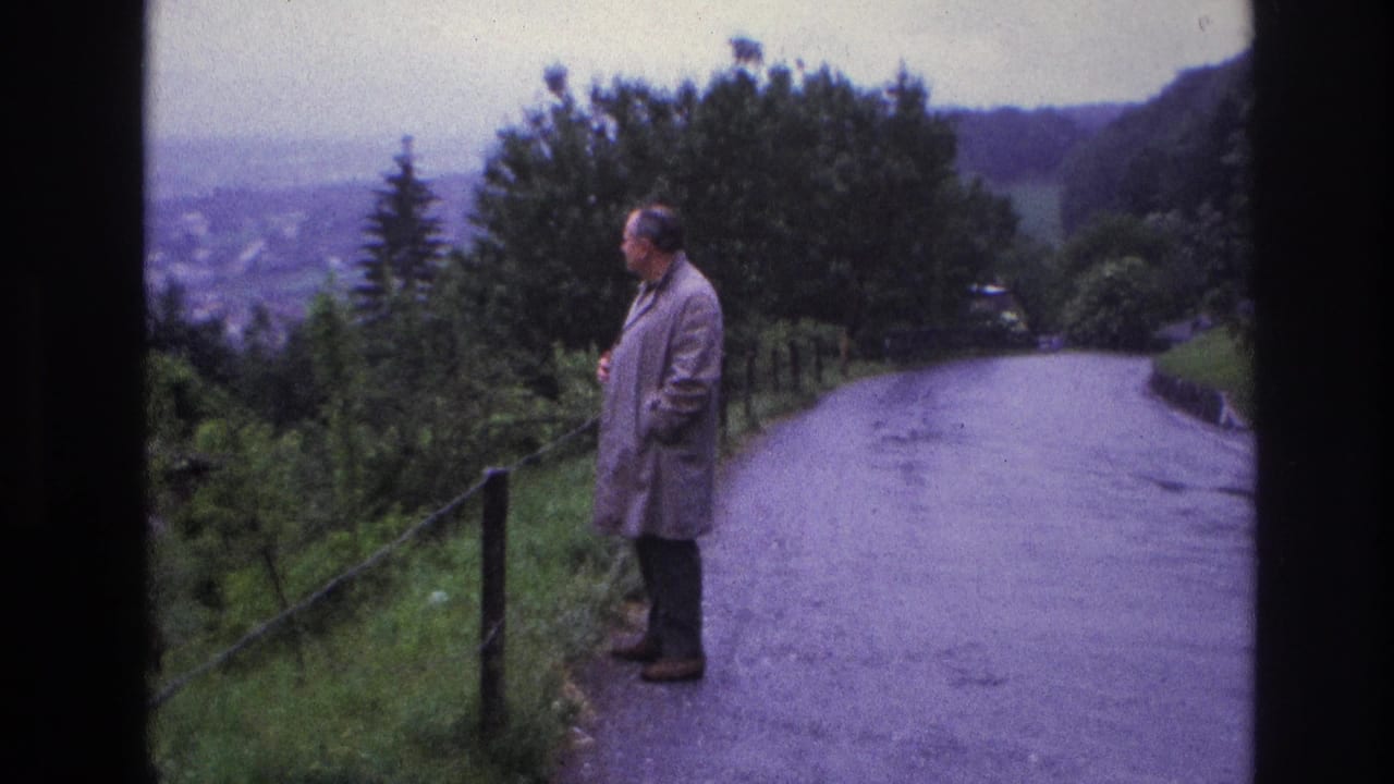 Man in grey raincoat pauses on a curved hillside road, France, circa 1967; a town is faintly visible in the valley below overcast skies.