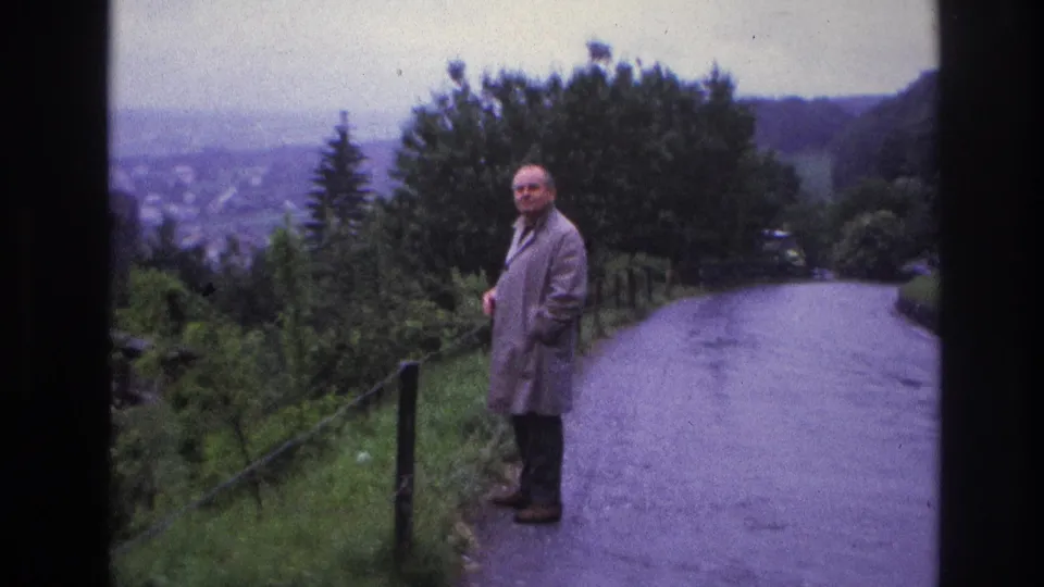 Man in grey raincoat turns toward camera on a wet hillside road, France, circa 1967; wooded hillside and broad valley spread behind him.