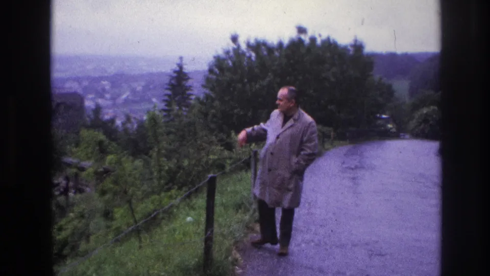Man in grey raincoat leans on a fence post and gestures toward the valley, France, circa 1967; distant town and overcast sky visible in background.