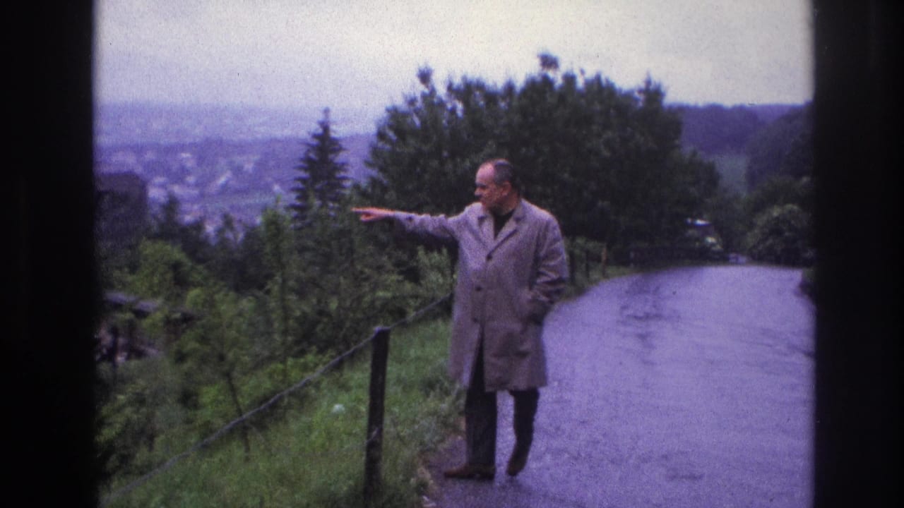 Man in light trench coat pointing along a winding hilltop road, France circa 1967; distant town panorama and wooden fence posts visible behind him.
