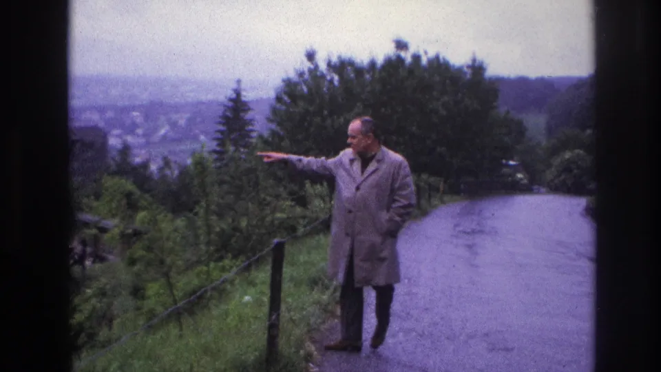 Man in light trench coat pointing along a winding hilltop road, France circa 1967; distant town panorama and wooden fence posts visible behind him.