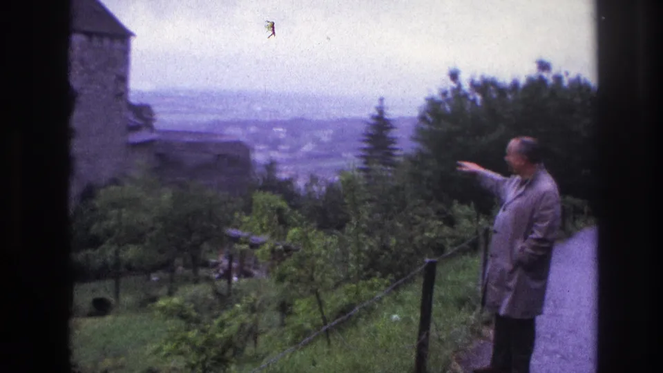 Man pointing beside a stone castle featuring a cylindrical conical-roofed tower on a wooded hillside, France circa 1967; panoramic town visible below.