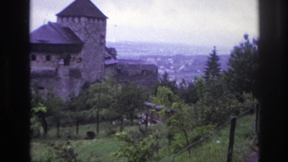 Medieval stone castle with cylindrical conical-roofed tower prominently framed; man in trench coat at right, large town panorama below, France circa 1967.