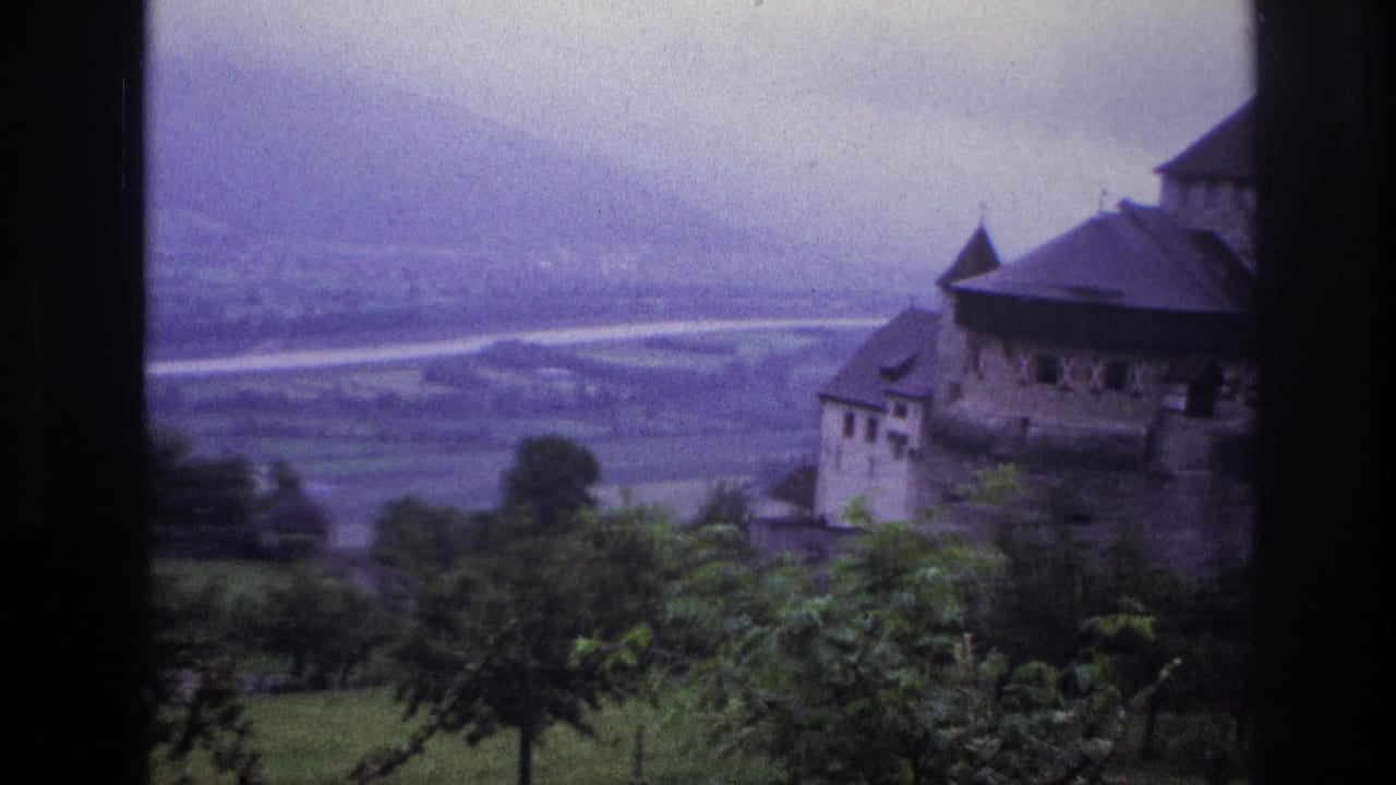 Same castle from slightly adjusted angle, valley and mountains in background, purple color shift on aged 8mm film.