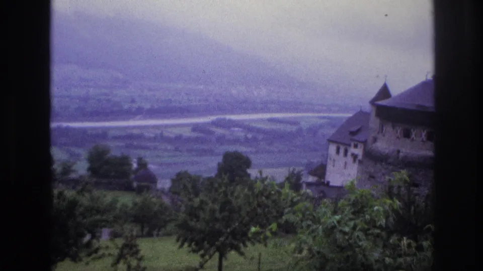 8mm home movie, France circa 1967, castle mid-frame after pan, more foreground tree canopy visible, broad flat valley with light-toned road or river beyond.
