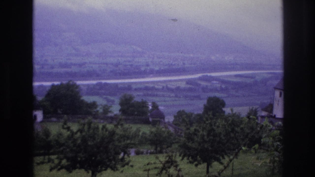8mm home movie frame, France 1967 — panoramic valley view through a window, with orchard trees, white farm buildings, and a river visible in the hazy distance.