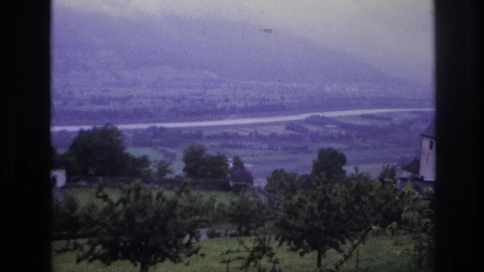 8mm home movie frame, France 1967 — panoramic valley view through a window, with orchard trees, white farm buildings, and a river visible in the hazy distance.