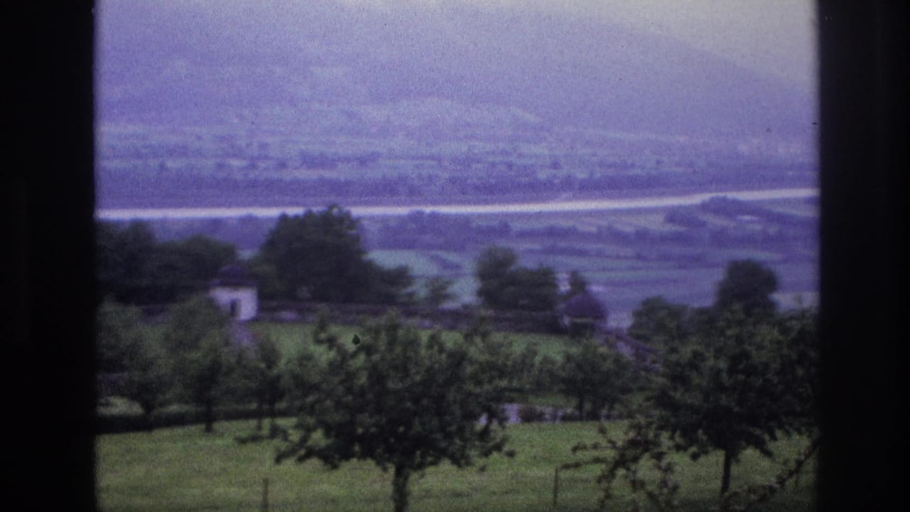 8mm home movie, France 1967 — valley view with white farm buildings and orchard trees; hazy mountain ridge in background, purple cast from aged reversal stock.