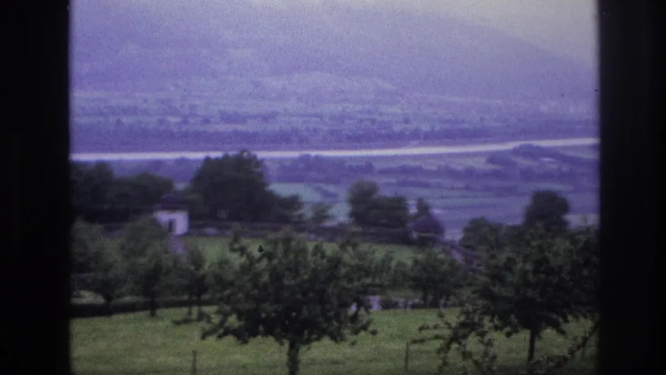 8mm home movie, France 1967 — valley view with white farm buildings and orchard trees; hazy mountain ridge in background, purple cast from aged reversal stock.