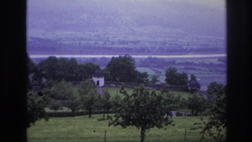 8mm home movie, France 1967 — valley view with a small white building left of frame, fruit trees in foreground, and broad valley floor stretching toward distant mountains.