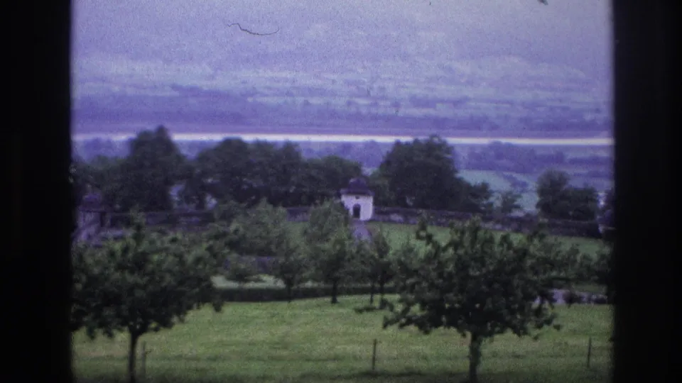 8mm home movie, France 1967 — final frame showing white building at right, orchard trees in foreground, and broad valley with pale river band under a hazy sky.