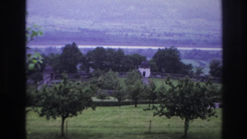 8mm home movie frame from France, circa 1967: summer landscape overlook with foreground trees, a white-roofed structure, and a wide valley under a pale overcast sky.