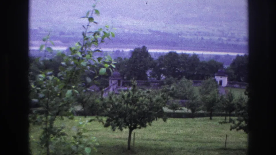 8mm home movie, France circa 1967: camera reframes to reveal a stone-walled terrace or garden structure in the midground; white-roofed building and hazy valley remain visible.