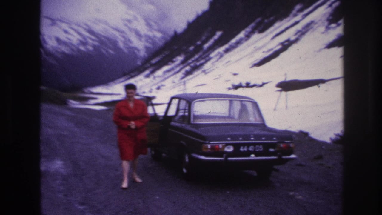 Woman in red dress near dark four-door sedan on alpine gravel road, France, circa 1967; steep snow-covered slopes and rocky peaks in background.