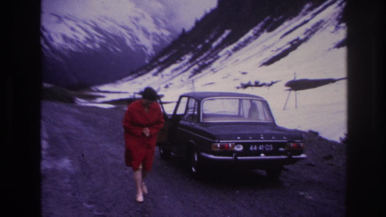 Woman in red dress turning beside dark 1960s sedan on gravel mountain road, snow-covered alpine peaks visible behind.