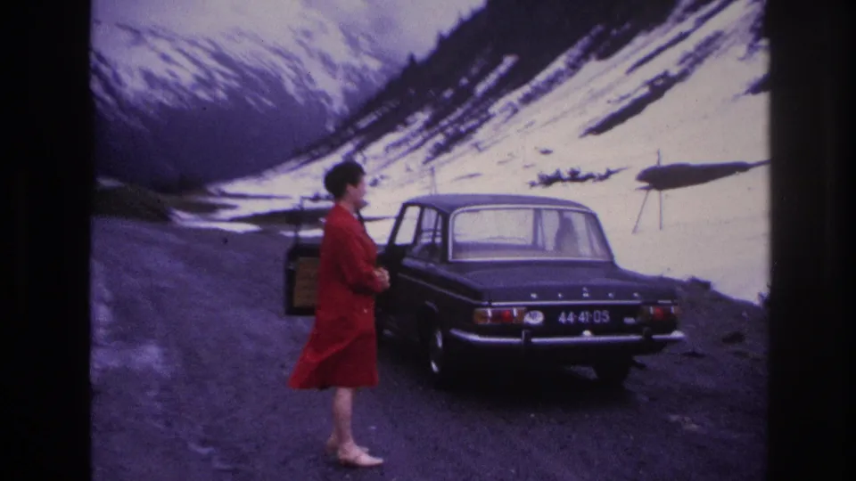 Woman in red dress at open car door of 1960s dark sedan on alpine mountain road, France, circa 1967; snow-covered peaks fill the background.