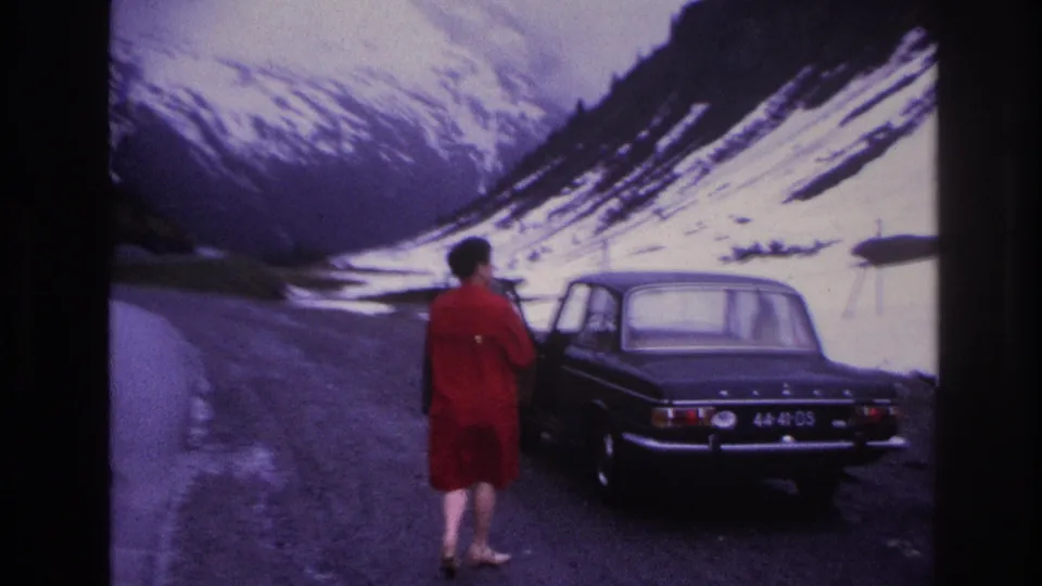 Woman in red dress from behind beside dark four-door sedan on gravel alpine road, French Alps, circa 1967; steep snow-covered peaks behind.