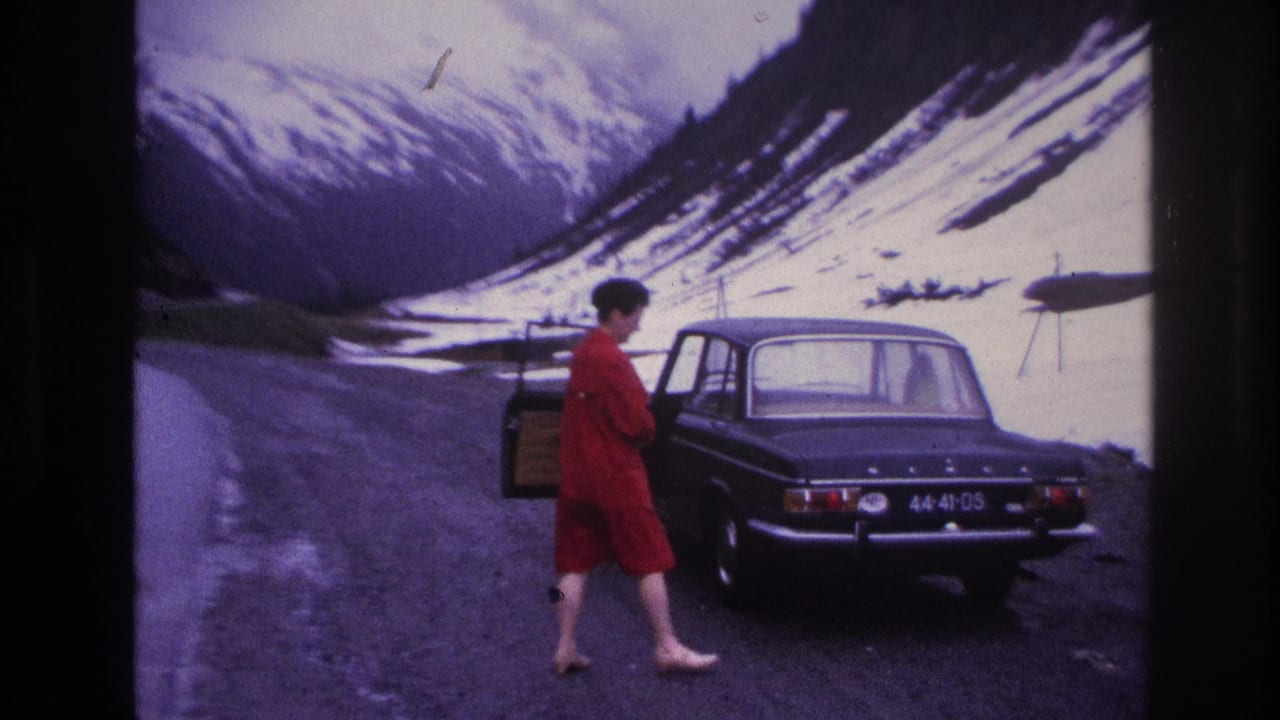 8mm film still, France c.1967 — barefoot woman in red dress approaching the open rear door of a dark European sedan on a high mountain road, snow-covered slopes rising steeply behind.