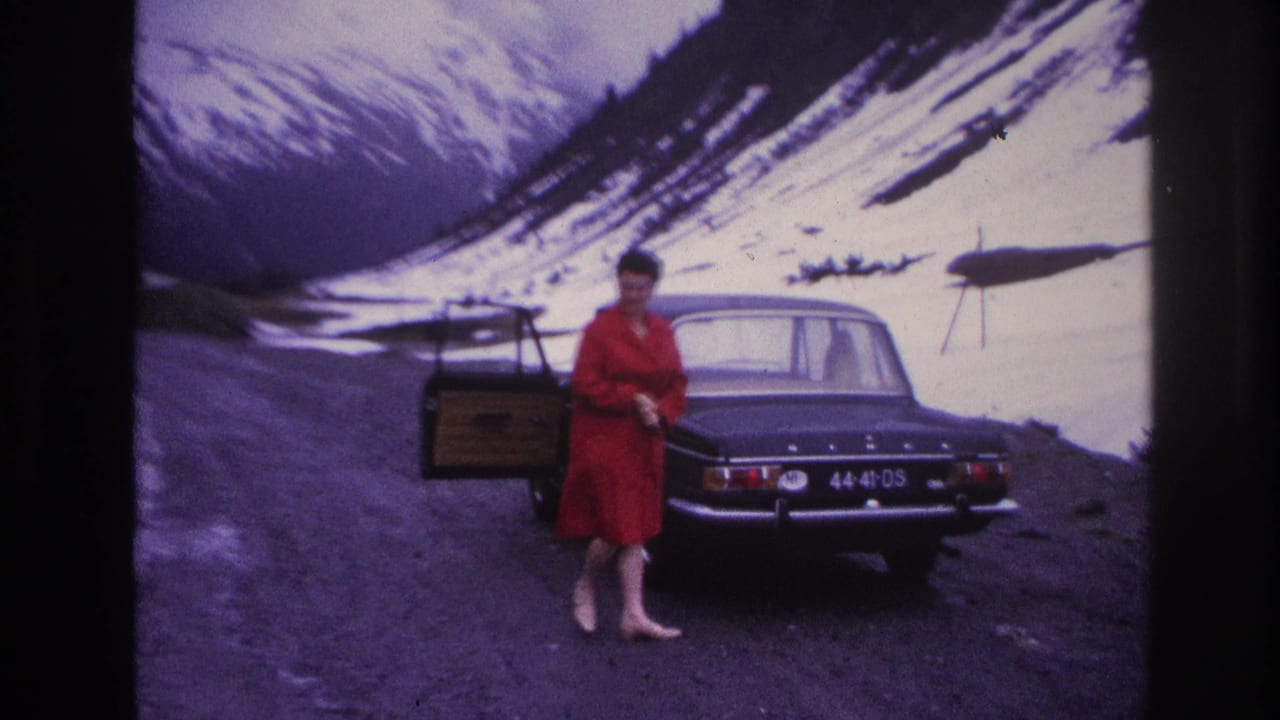Woman in red dress standing beside open rear door of dark sedan on mountain road, steep snow slopes filling background.